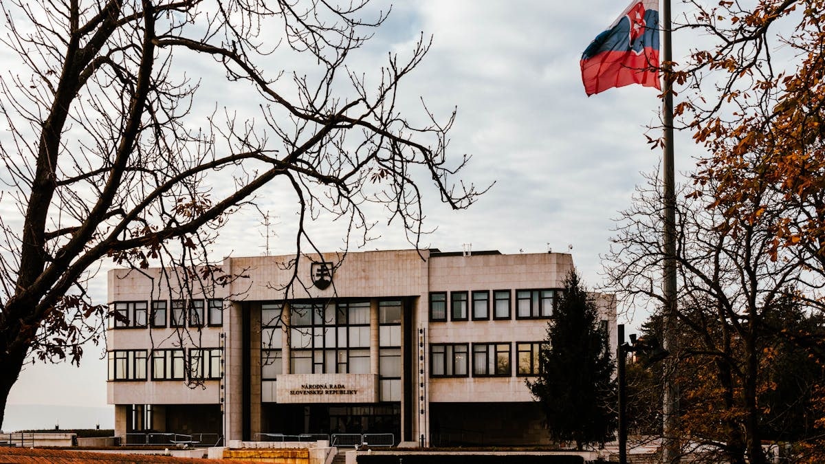 A view of the Slovak Parliament building surrounded by autumn trees with the national flag waving under an overcast sky.